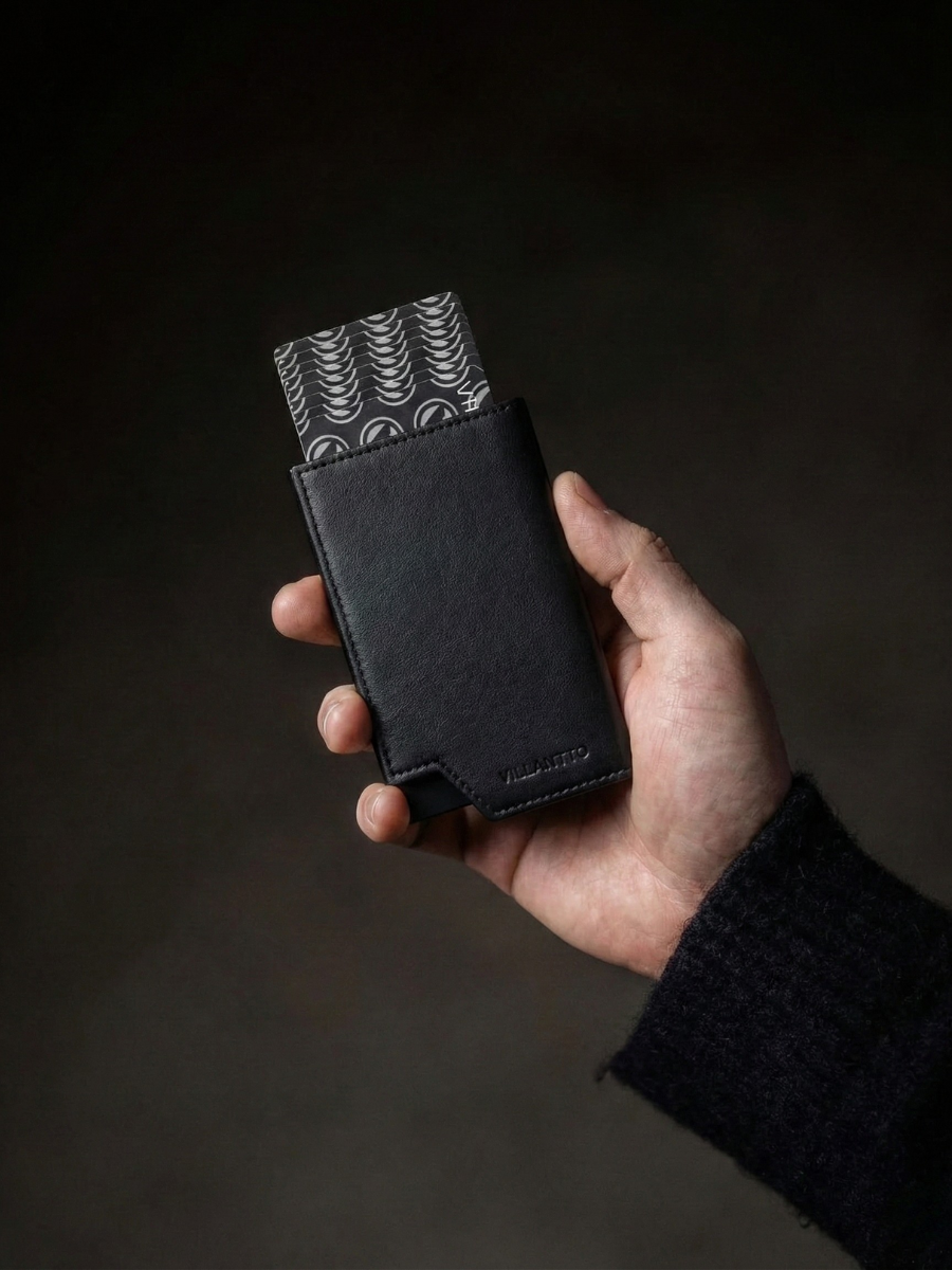 Hand holding a black leather wallet with patterned flap against a dark background